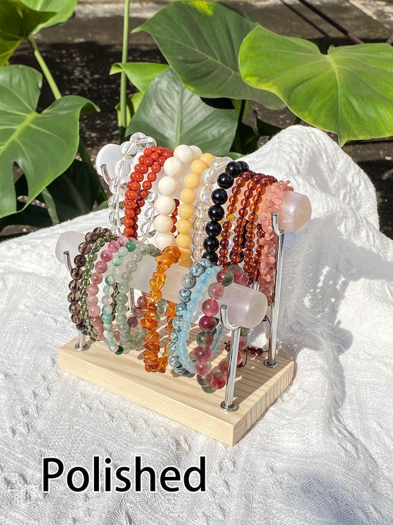 K&ouml;nnte beinhalten: Verschiedene Armb&auml;nder aus Edelsteinen in Rot, Wei&szlig;, Gelb, Schwarz und Rosa. Die Armb&auml;nder sind auf einem Holzst&auml;nder mit Metallst&uuml;tzen angeordnet. Das Wort "Polished" ist am unteren Bildrand zu sehen.