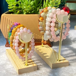 Peut inclure: Présentation de bracelets de perles de différentes couleurs, notamment rose, orange et violet, présentés sur des présentoirs en bois avec des supports métalliques dorés. Les bracelets sont faits de pierres précieuses et de perles. Une plante succulente est en arrière-plan.