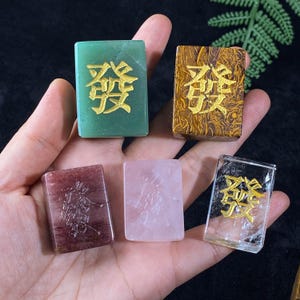 Op de afbeelding: Een hand houdt vijf rechthoekige, gepolijste edelsteen tegels vast. De tegels zijn groen, bruin, rood, roze en helder. Elke tegel heeft gouden Chinese karakters. De edelstenen zijn glad en reflecteren licht.