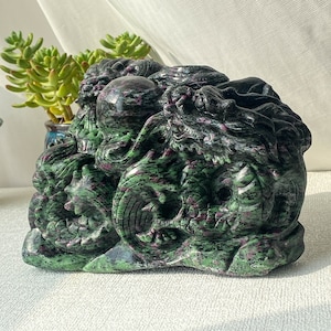 Op de afbeelding: Een groene en roze jade-beeldhouwwerk van een draak. De draak is ingewikkeld gedetailleerd en heeft een wervelende, organische vorm.