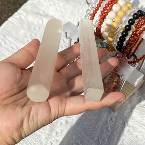 K&ouml;nnte beinhalten: Zwei durchscheinende Selenit-Zauberst&auml;be, einer abgerundet und einer quadratisch, werden in einer Hand gehalten. Eine Vielzahl von Perlenarmb&auml;ndern in Rot-, Wei&szlig;-, Gelb-, Schwarz- und Braunt&ouml;nen sind im Hintergrund. Die Zauberst&auml;be sind hell.