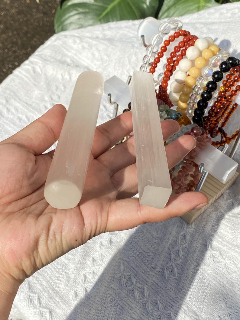 K&ouml;nnte beinhalten: Zwei durchscheinende Selenit-Zauberst&auml;be, einer abgerundet und einer quadratisch, werden in einer Hand gehalten. Eine Vielzahl von Perlenarmb&auml;ndern in Rot-, Wei&szlig;-, Gelb-, Schwarz- und Braunt&ouml;nen sind im Hintergrund. Die Zauberst&auml;be sind hell.