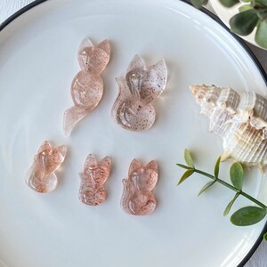 以下が含まれることがあります： 白い皿の上に、透き通ったピンクと茶色の小さな狐の彫刻が5つ置かれています。狐は水晶でできています。