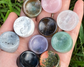 Op de afbeelding: Een verzameling van tien gepolijste, ronde edelstenen in verschillende kleuren en patronen. De stenen omvatten tinten roze, groen, bruin, zwart en wit. De edelstenen worden in een hand gehouden tegen een achtergrond van groen gebladerte.