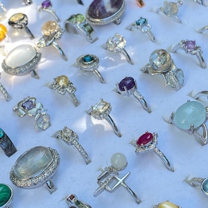 May include: A collection of assorted metal rings featuring various gemstones including amethyst, moonstone, ruby, and opal. The rings are arranged in rows on a white display tray, showcasing diverse settings, band designs, and colorful cabochon stones.