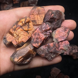 May include: A selection of raw labradorite stones, displaying a blend of orange, red, and purple hues. The stones have a rugged, textured surface and are held in a hand, emphasizing their natural beauty and unique mineral composition.