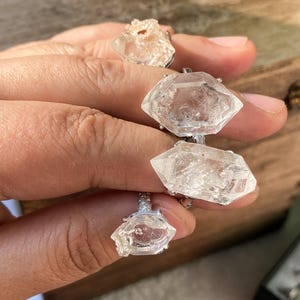 以下が含まれることがあります： 宝石リングのコレクション。クリアでファセットカットされた水晶と、銀色のバンドにセットされたラフカットの明るい色の石が特徴です。リングは木製の表面に展示されています。