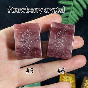 Pu&ograve; includere: Due cristalli di fragola rettangolari, numerati #5 e #6, sono tenuti in una mano. I cristalli sono di un colore rosso intenso con motivi intricati. Il testo "Strawberry crystal" &egrave; nella parte superiore dell'immagine.