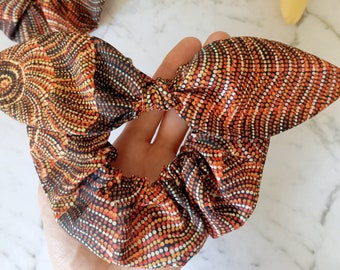 May include: A brown, orange and yellow patterned hair scrunchie with a black background. The scrunchie is made of fabric and has a gathered design.