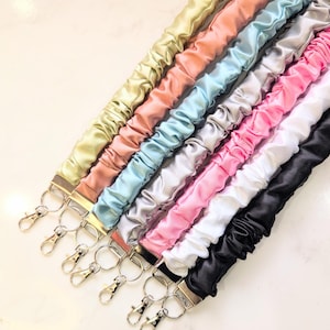 May include: A collection of satin lanyards in various colors, including gold, peach, light blue, silver, pink, white, and black. Each lanyard has a metal clasp and ring for attaching keys or an ID badge. The lanyards are arranged diagonally.