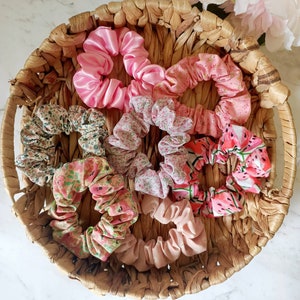 May include: A wicker basket filled with eight colorful scrunchies. The scrunchies are made of fabric with floral and watermelon prints. There are also two solid pink scrunchies.