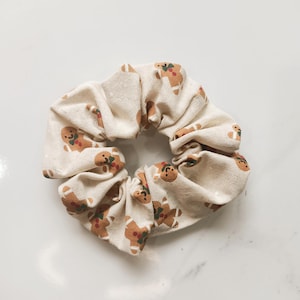 May include: A cream-colored scrunchie with a repeating pattern of gingerbread men. The gingerbread men are brown with red and green accents. The scrunchie is made of a soft fabric and is suitable for all hair types.
