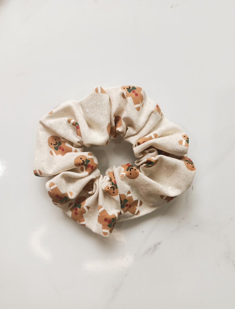May include: A cream-colored scrunchie with a repeating pattern of gingerbread men. The gingerbread men are brown with red and green accents. The scrunchie is made of a soft fabric and is suitable for all hair types.