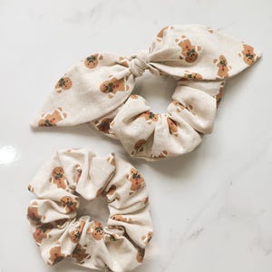 May include: Two beige scrunchies with a repeating gingerbread man pattern. One scrunchie has a bow, and the other is a standard scrunchie. The gingerbread men have brown icing and green accents. The scrunchies are made of a soft fabric.