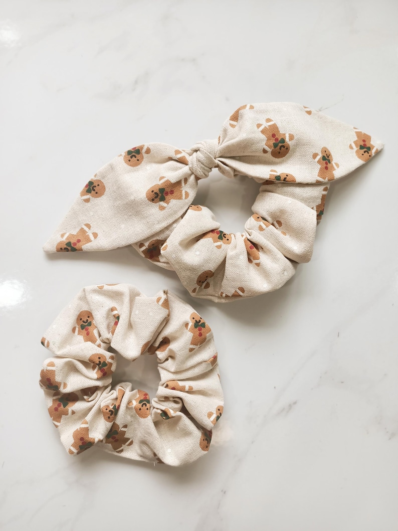 May include: Two beige scrunchies with a repeating gingerbread man pattern. One scrunchie has a bow, and the other is a standard scrunchie. The gingerbread men have brown icing and green accents. The scrunchies are made of a soft fabric.