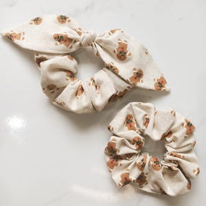 May include: Two beige scrunchies with a repeating gingerbread man pattern. One scrunchie has a bow, and the other is a standard scrunchie. The gingerbread men are brown with red and green accents. The background is a light beige.