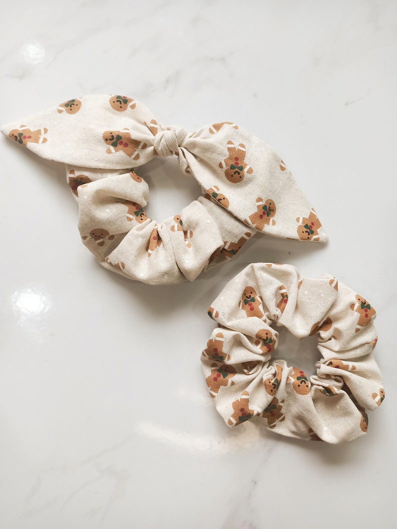 May include: Two beige scrunchies with a repeating gingerbread man pattern. One scrunchie has a bow, and the other is a standard scrunchie. The gingerbread men are brown with red and green accents. The background is a light beige.
