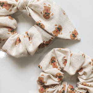 May include: Two beige scrunchies with a repeating pattern of gingerbread men. The scrunchies have a bow detail. The gingerbread men are brown with green bow ties and red accents. The background is a light beige color.