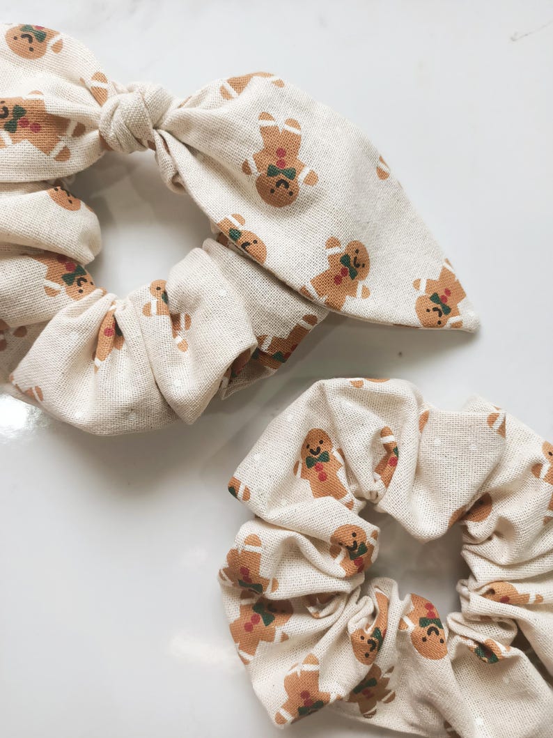 May include: Two beige scrunchies with a repeating pattern of gingerbread men. The scrunchies have a bow detail. The gingerbread men are brown with green bow ties and red accents. The background is a light beige color.