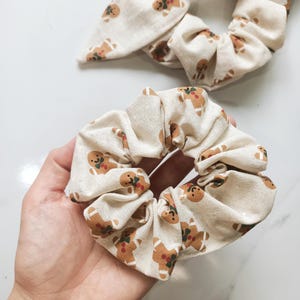 May include: Two beige fabric scrunchies with a repeating gingerbread man pattern. One scrunchie is a standard circular design, and the other has a bow. The gingerbread men are brown with red and green accents.