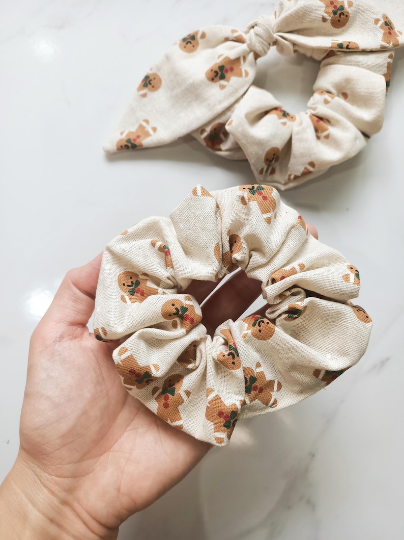 May include: Two beige fabric scrunchies with a repeating gingerbread man pattern. One scrunchie is a standard circular design, and the other has a bow. The gingerbread men are brown with red and green accents.