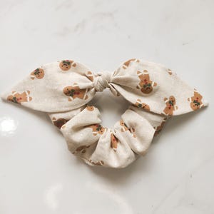 May include: A beige scrunchie with a bow, adorned with a repeating pattern of gingerbread men. The gingerbread men are brown with red and green accents. The scrunchie is a festive hair accessory.