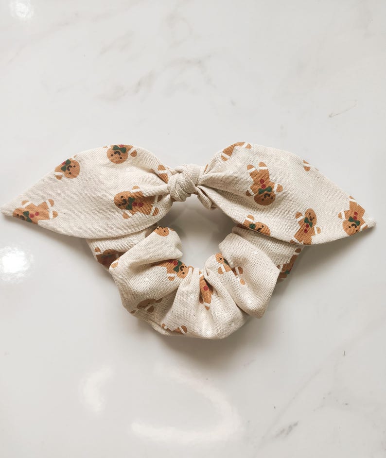 May include: A beige scrunchie with a bow, adorned with a repeating pattern of gingerbread men. The gingerbread men are brown with red and green accents. The scrunchie is a festive hair accessory.