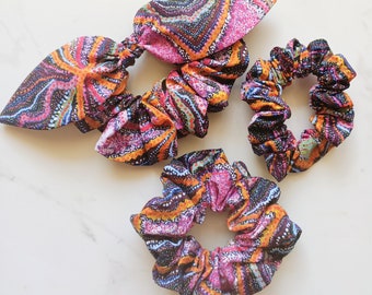 May include: Three colourful hair scrunchies with an abstract pattern in shades of pink, orange, brown, and blue. The scrunchies are made of fabric and have a gathered, ruffled look.