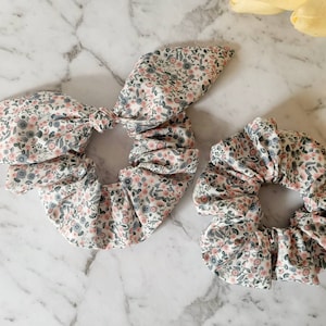 May include: Two floral print scrunchies, one with a bow, in shades of pink, white, and gray.