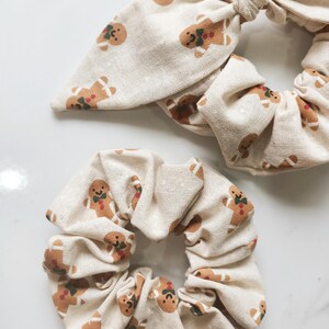 May include: Two beige scrunchies with a repeating gingerbread man pattern. One scrunchie has a bow detail. The gingerbread men have brown bodies, green bow ties, and red accents. The background is a light beige.