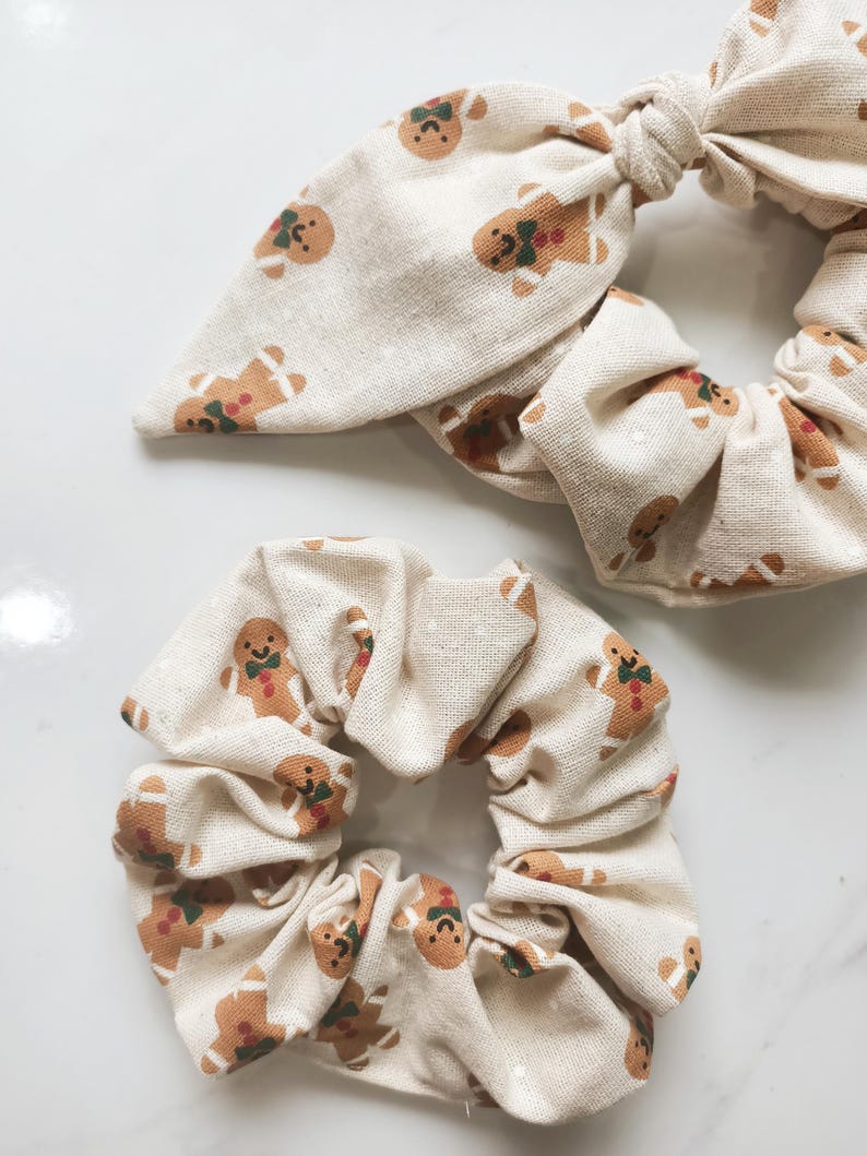 May include: Two beige scrunchies with a repeating gingerbread man pattern. One scrunchie has a bow detail. The gingerbread men have brown bodies, green bow ties, and red accents. The background is a light beige.