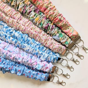 May include: A collection of six fabric lanyards with floral patterns in various colors, including pink, blue, and green. Each lanyard has a metal clasp and key ring. The fabric is gathered, creating a textured appearance.