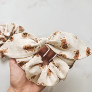 May include: A beige scrunchie with a bow, patterned with gingerbread men. The scrunchie is made of fabric and is tied in a bow shape. The gingerbread men are brown with green and red accents. A second scrunchie is visible in the background.