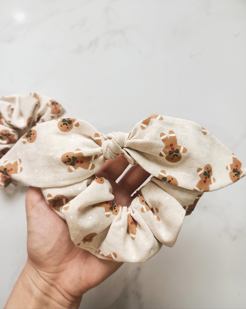 May include: A beige scrunchie with a bow, patterned with gingerbread men. The scrunchie is made of fabric and is tied in a bow shape. The gingerbread men are brown with green and red accents. A second scrunchie is visible in the background.