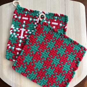 May include: Two woven potholders in red, green, and white, with wooden ring loops for hanging. One features a snowflake design, the other a geometric pattern. They rest on a light-colored wooden surface, suitable for kitchen use.