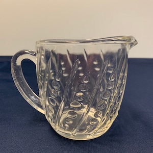 May include: A clear glass creamer with a handle and a spout. The creamer has a textured pattern of diagonal lines and circles. The creamer is sitting on a dark blue surface.