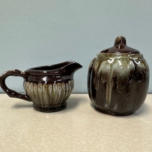 May include: A brown and white ceramic creamer and sugar set with a drip glaze. The creamer has a fluted design and a handle. The sugar bowl has a lid.