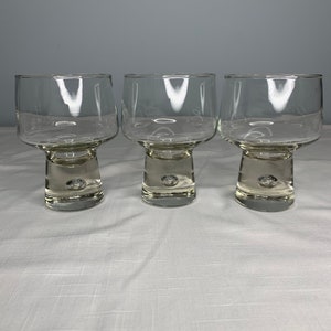 May include: Three clear, stemware glasses with a modern design. Each glass features a wide, square-shaped bowl atop a short, cylindrical stem with a small bubble detail. The glasses are arranged on a white surface.
