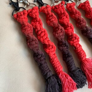 May include: A collection of macrame keychains featuring a spiral knot pattern in an ombre color gradient that transitions from bright red to dark brown. Each keychain is finished with a frayed tassel at the bottom and attached to a metal lobster clasp.