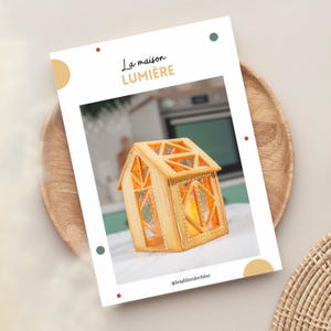 Peut inclure: Une maison en pain d'épices en forme de lanterne avec une lueur jaune à l'intérieur. La maison est décorée de glaçage blanc et le texte "La maison LUMIÈRE" est écrit sur le devant. L'image est légendée avec "@letablierdechloe".