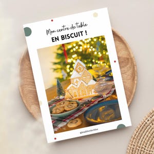 Könnte beinhalten: Ein weißes Papier mit dem Text "Mon centre de table EN BISCUIT!" zeigt ein Lebkuchenhaus-Mittelstück auf einem Tisch. Der Tisch ist mit Keksen, Getränken und einem Weihnachtsbaum im Hintergrund gedeckt. Das Papier liegt auf einem Holztablett.