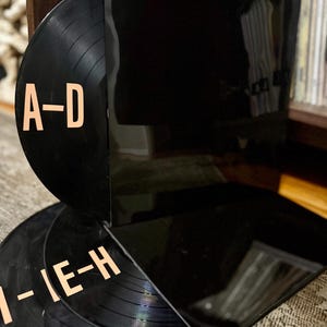 Puede incluir: Una pila de discos de vinilo negros con letras de color naranja claro. Los discos están etiquetados como "A-D" e "I-IE-H". Una funda de disco negra está parcialmente abierta, revelando los discos. La imagen está tomada en interiores.
