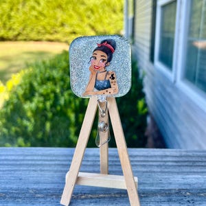 May include: A silver glitter square badge holder with a cartoon image of a woman with a red headband and a tattoo on her arm. The badge holder is on a small wooden easel.