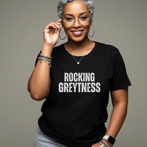May include: A woman with short, silver-grey hair is wearing a black t-shirt with the text "ROCKING GREYTYNESS" in white. She is wearing round glasses and a silver necklace. She is smiling and looking at the camera.