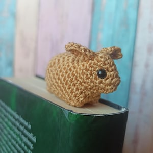May include: A small, crocheted bunny in a light brown color with black eyes. The bunny is sitting on top of a green book. The bunny's ears are upright, and the body is round. The bunny is a handmade item.