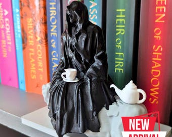 May include: A black figurine of a hooded figure sitting on a white base, holding a small white teacup and saucer. A white teapot is on the base to the right of the figure. The figurine is on a shelf with a row of books behind it. The books have titles like "Throne of Glass" and "Queen of Shadows". The text "NEW ARRIVAL" is on a red sticker in the lower right corner of the image.