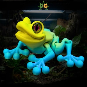 May include: A 3D-printed frog sculpture with a vibrant color gradient. The frog features a yellow head, transitioning to blue legs and feet. The eyes are detailed with purple and black accents. The sculpture is set against a backdrop of greenery.