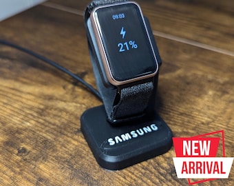 May include: A black Samsung smartwatch charging on a black charging stand. The watch face shows the time 09:03 and a battery icon with 21%.