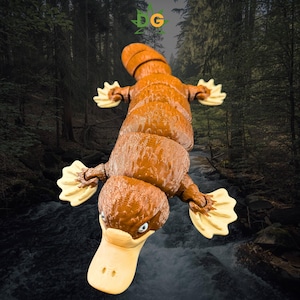 May include: A brown and tan platypus figurine with a textured body and webbed feet. The figurine has a duck-like bill and small, blue eyes. The background features a forest scene with a flowing river.