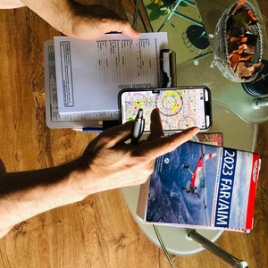 Puede incluir: Una persona está revisando un documento de papel y un teléfono móvil con un mapa en la pantalla. Un libro titulado "2023 FAR/AIM" está sobre una mesa de vidrio.
